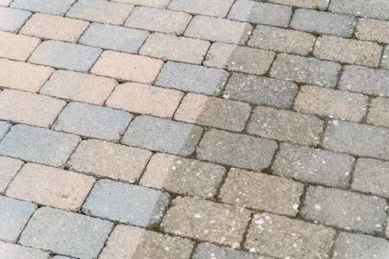 Bild på uppfartens stenläggning med rätvinkliga ljusgrå och rosa betongstenar, vissa delar täckta av mossa och vita beläggningar. Nanostone utför altantvätt, stentvätt och takbehandling för att återställa ytors färg och fogars skick med miljöanpassade metoder i tryggt och säkert utförande.