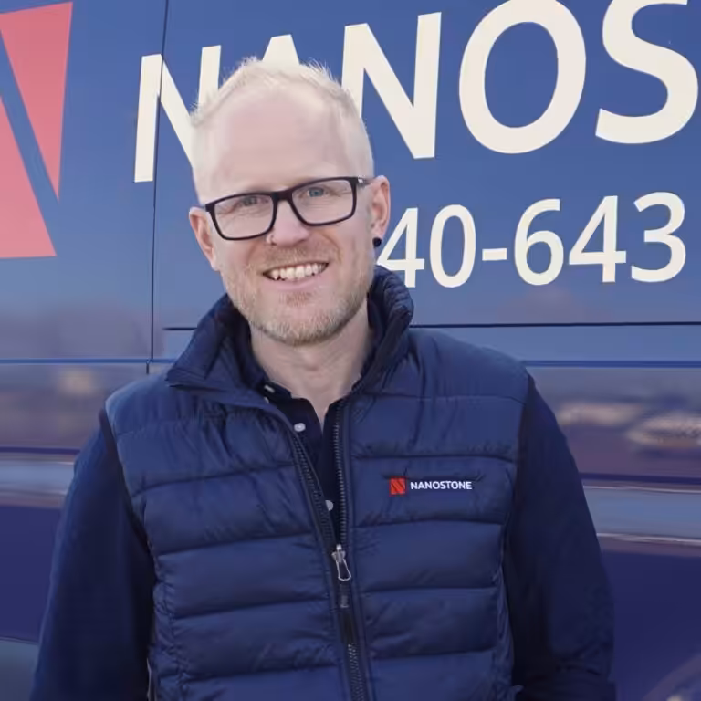 Man i 40–50-årsåldern med glasögon och kort ljust hår, klädd i en mörk Nanostone‑väst, ler framför en blå företagsbil med Nanostone‑logotyp och telefonnummer. Bilden visar företagets profil och tjänster: altantvätt, stentvätt och takbehandling utförda tryggt av erfaren personal med många års erfarenhet.