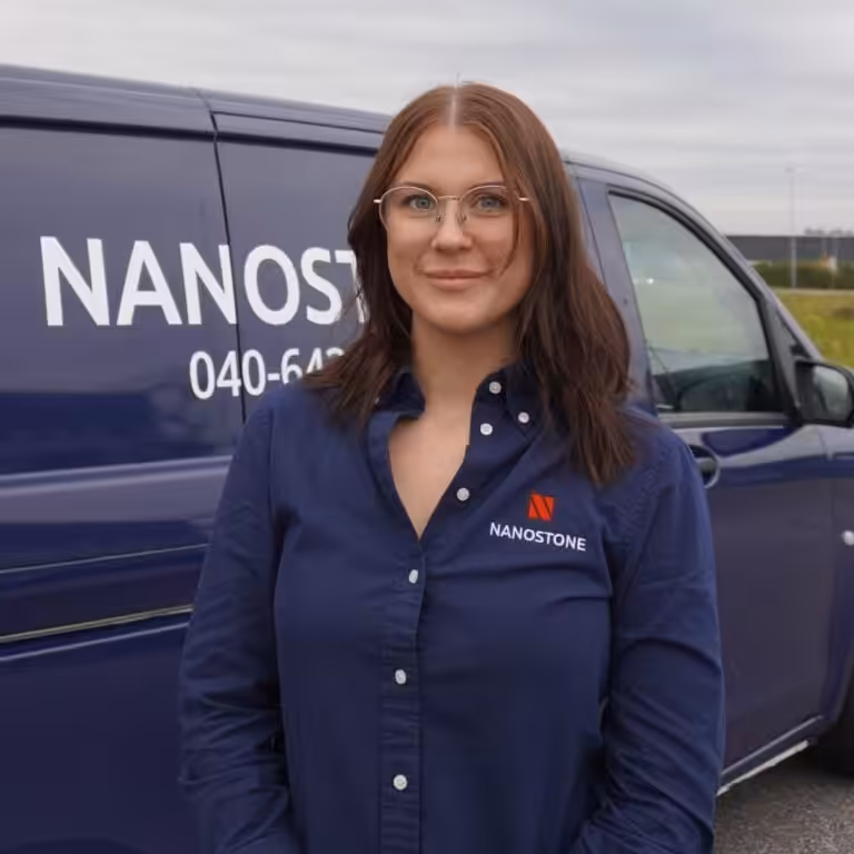 Kvinna i blå Nanostone-skjorta och uniform står leende med glasögon framför företagets blå transportbil med tydlig logotyp. Bilden visar en professionell, engagerad och välvårdad medarbetare, redo för uppdrag, specialiserad på altantvätt, stentvätt och takbehandling, med lång erfarenhet och trygg serviceprofil.