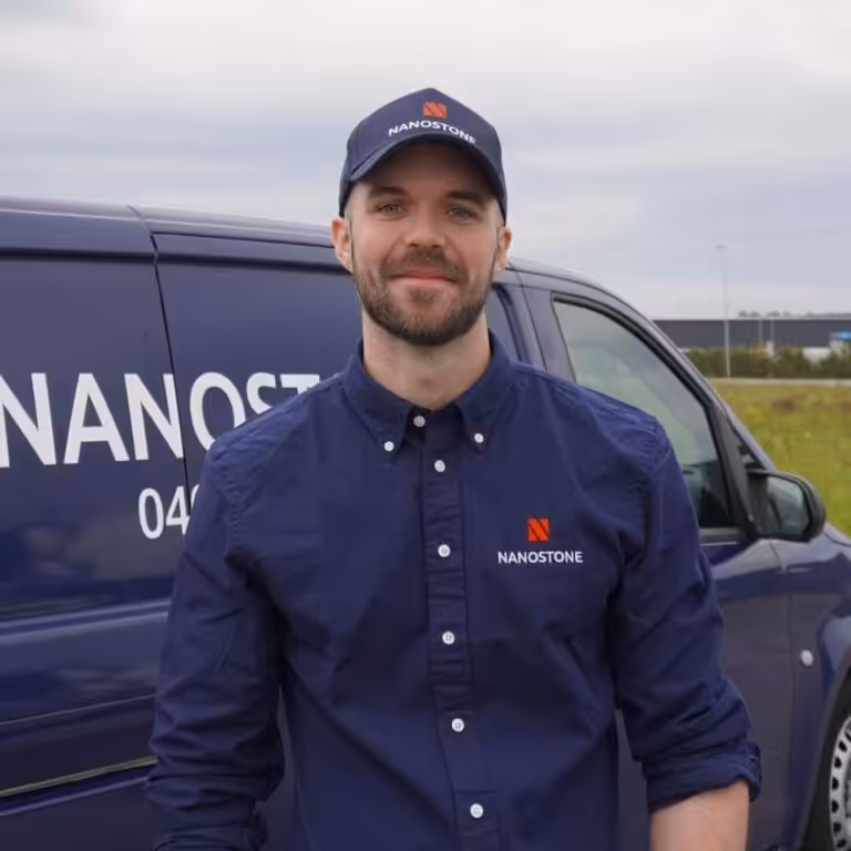 En leende man i Nanostone-keps och företagsblå skjorta med uppkavlade ärmar står framför en blå Nanostone-arbetsbil, utomhus vid en gräsplan och industribyggnader. Porträttet visar en skäggig, erfaren tekniker med företagets logotyp synlig, redo för altantvätt, stentvätt och takbehandling, professionell och serviceinriktad.