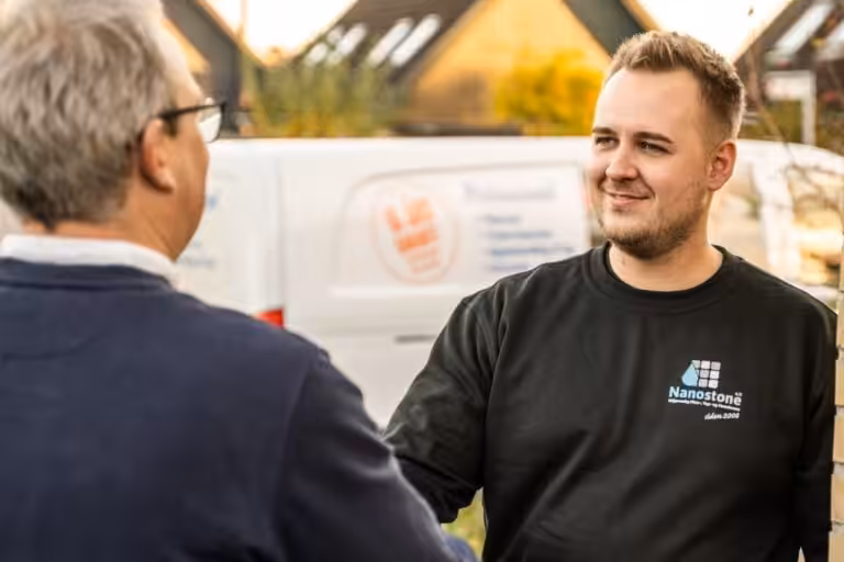 Två män i förgrunden, en i Nanostone-tröja som möter och skakar hand med en kund utanför ett bostadshus; vit företagsbil och bostadstak syns i bakgrunden. Bilden kommunicerar professionell, erfaren och trygg service från Nanostone för altantvätt, stentvätt och takbehandling i bostadsområden.