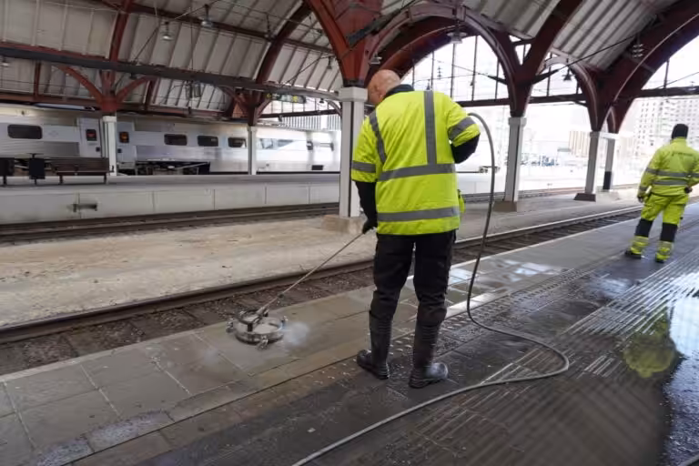 Nanostone-team i varselkläder högtryckstvättar perrongplattor vid en stationsplattform under stora röda valvbågar; en operatör hanterar cirkulärt markrengöringsverktyg och slang medan kollega står vid rälsen. Bilden visar välutrustad, erfaren personal som utför altantvätt, stentvätt och takbehandling på offentliga ytor med säkerhetsutrustning och noggrann teknik.