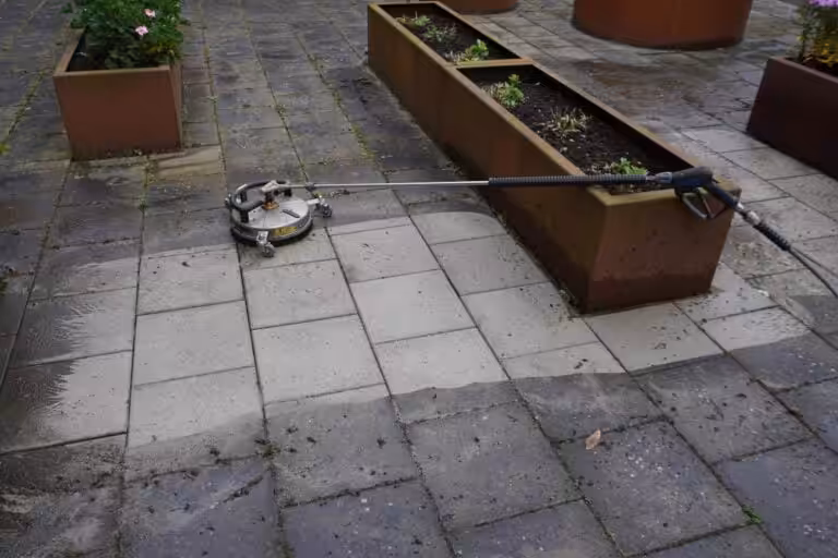 Bild visar Nanostone:s yrkesmässiga rengöring av utomhusplattor med roterande högtrycksyta och lanskoppling, där en mörk, algig stenyta tydligt kontrasterar mot nyligen rengjorda, ljusare plattor. Planteringslådor och blöta ytor syns samt rengöringsslang och hjul. Tjänster: altantvatt, stentvatt, takbehandling. Erfaren personal ger en professionell finish vid utomhusrengöring.