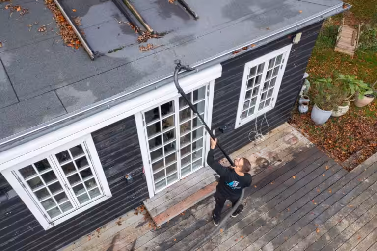 Nanostone personal använder lång ställd stav och sugslang för takbehandling av ett plant tak och rensning vid takränna på en svart träfasad. Bilden från ovan visar fönsterpartier, löv på taket och en arbetare på trätrall — professionell altantvätt, stentvätt och takbehandling.