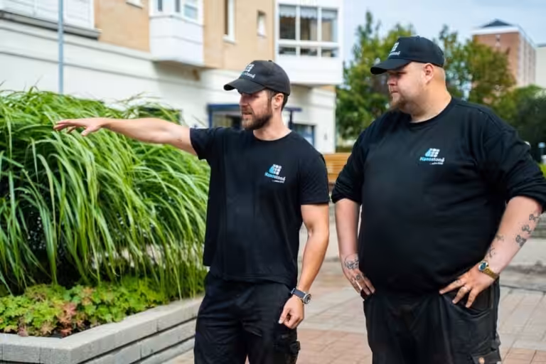 Två Nanostone-medarbetare i svarta arbetskläder och kepsar står utomhus framför en bostadsfasad och planteringar; den ena pekar mot området medan den andre lyssnar. Bilden illustrerar Nanostones arbete med altantvätt, stentvätt och takbehandling i en professionell, erfaren och säker arbetsmiljö med fokus på kvalitet.