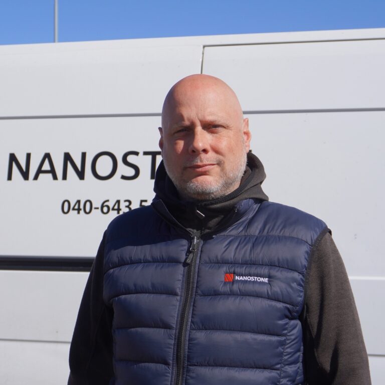 En uniformerad man i Nanostone-väst står framför en vit servicebil med företagsnamnet synligt; professionellt porträtt i dagsljus. Vadderad mörkblå väst med logotyp och svart långärmad tröja syns. Bilden signalerar tjänster för altantvätt, stentvätt och takbehandling från ett erfaret rengöringsföretag specialister.