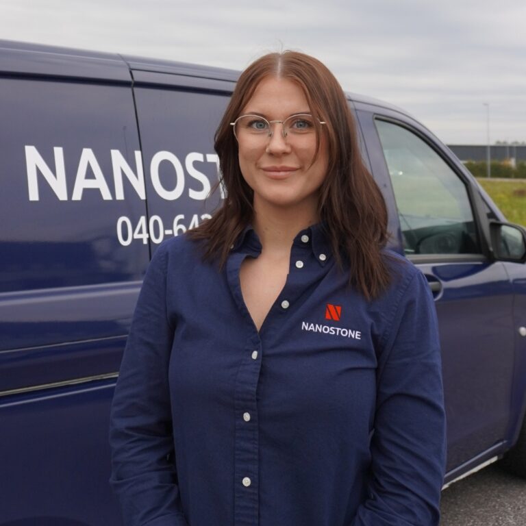 En kvinnlig Nanostone-medarbetare i mörkblå företagsblus med Nanostone-logotyp och knappar står leende framför en mörkblå servicebil utomhus med delvis synligt telefonnummer och företagsnamn. Hon har axellångt brunt hår och glasögon; bilden presenterar en professionell, erfaren profil för altantvätt, stentvätt och takbehandling.