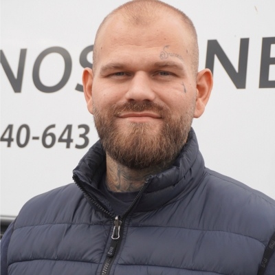 Porträtt av en man i mörk vadderad jacka med skägg och synliga ansikts- och nacktatueringar, stående framför en Nanostone-skylt på företagets fordon. Bilden förmedlar professionellt utomhusteam och företagets tjänster: altantvätt, stentvätt och takbehandling utförda med erfarenhet, modern utrustning och fokus på säkerhet.